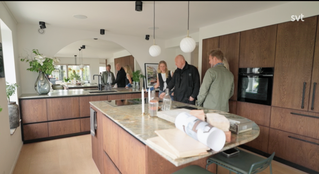 Stiligt - Fyra personer står och pratar i ett modernt kök med skåp i mörkt trä, en ö i marmor och pendellampor. Arkitektritningar och material ligger utspridda på bänken.