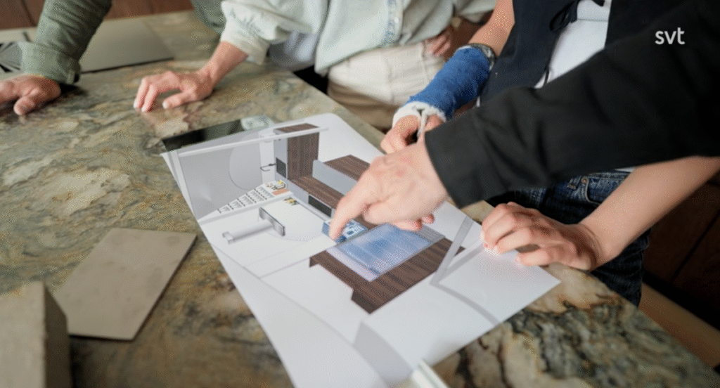 Stiligt - Tre personer tittar på och diskuterar en 3D-utskrift av en köksritning på en marmorbänkskiva; en person pekar på ritningen medan en annan har armen i blått gips.