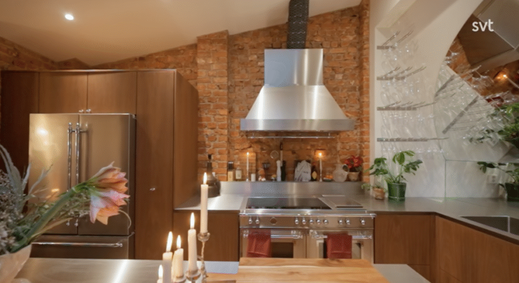 Stiligt - Ett modernt kök med synliga tegelväggar, vitvaror i rostfritt stål, en stor ugn, glashyllor med glasföremål och ett kylskåp. Stearinljus och växter ger en dekorativ touch.