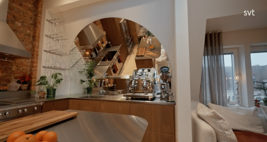 Stiligt - Modernt kök med bänkskivor i rostfritt stål, kaffemaskin, glasögon på hyllor och en stor rund spegel som speglar rummet; intill ett mysigt vardagsrum med fönster.