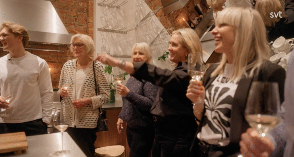 Stiligt - En grupp människor står i ett modernt kök, håller i vinglas och ler; en kvinna pekar mot något utanför kameran.