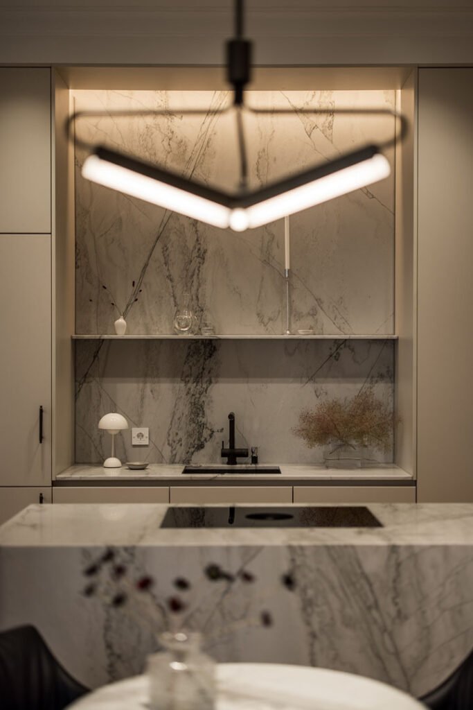 Stiligt - Modernt kök med stänkskydd och bänkskivor i marmor, minimalistiska hyllor, svart blandare, köksö av vacker natursten, inbyggd spishäll och en pendelarmatur i förgrunden.