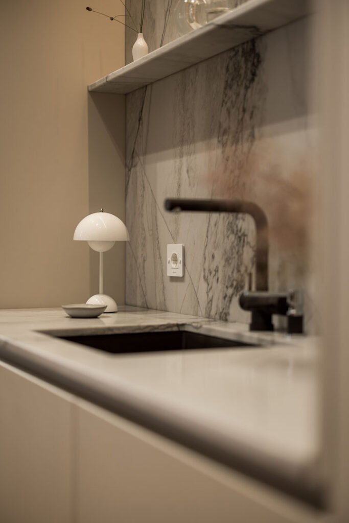 Stiligt - Minimalistisk köksbänk med marmorbacksplash, svart kran, vit bänkskiva, en modern vit bordslampa och en liten skål på bänken, vackert kompletterad med en köksö av vacker natursten.