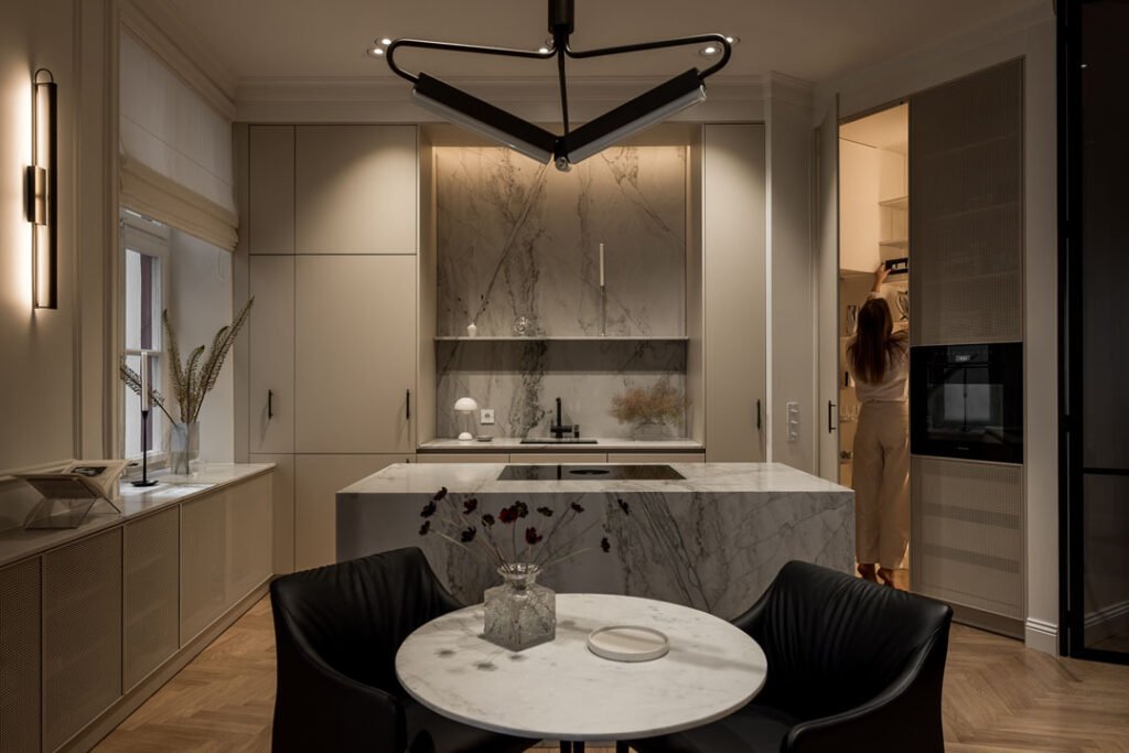 Stiligt - Modernt kök med marmorbänkskivor, köksö av vacker natursten, snyggt stänkskydd, runt matbord i förgrunden och en person som står vid en dörröppning i bakgrunden.