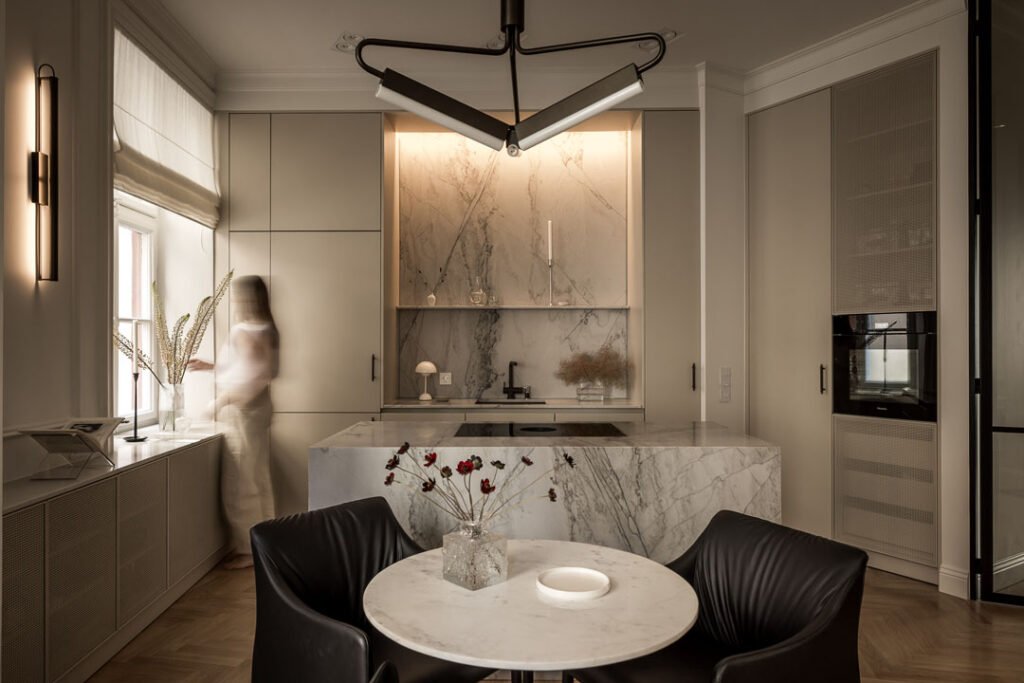 Stiligt - Modernt kök med marmorbänkskivor, köksö av vacker natursten, stänkskydd, runt matbord med svarta stolar och en person som står vid fönstret i dämpad belysning. Varför ett platsbyggt kök ofta är billigare än du tror på sikt: Smarta spartips!