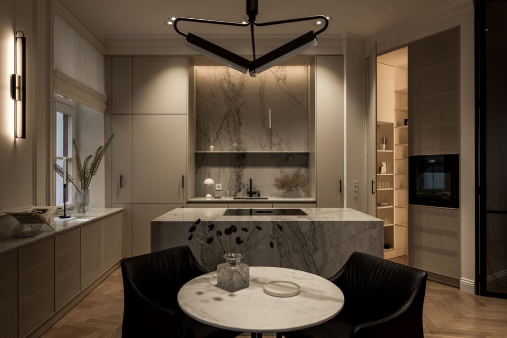 Stiligt - Modernt kök med köksö av vacker natursten med marmorbacksplash, runt matbord med två svarta stolar och minimalistisk belysning i ett svagt upplyst utrymme.