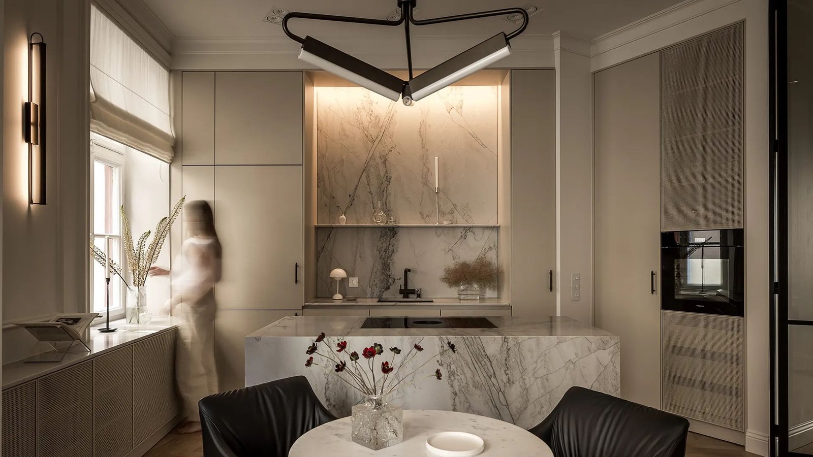 Stiligt - Ett modernt kök med bänkskivor och stänkskydd i marmor, beige köksskåp, en köksö i vacker natursten, ett runt matbord med torkade blommor och en suddig person som står vid ett fönster.
