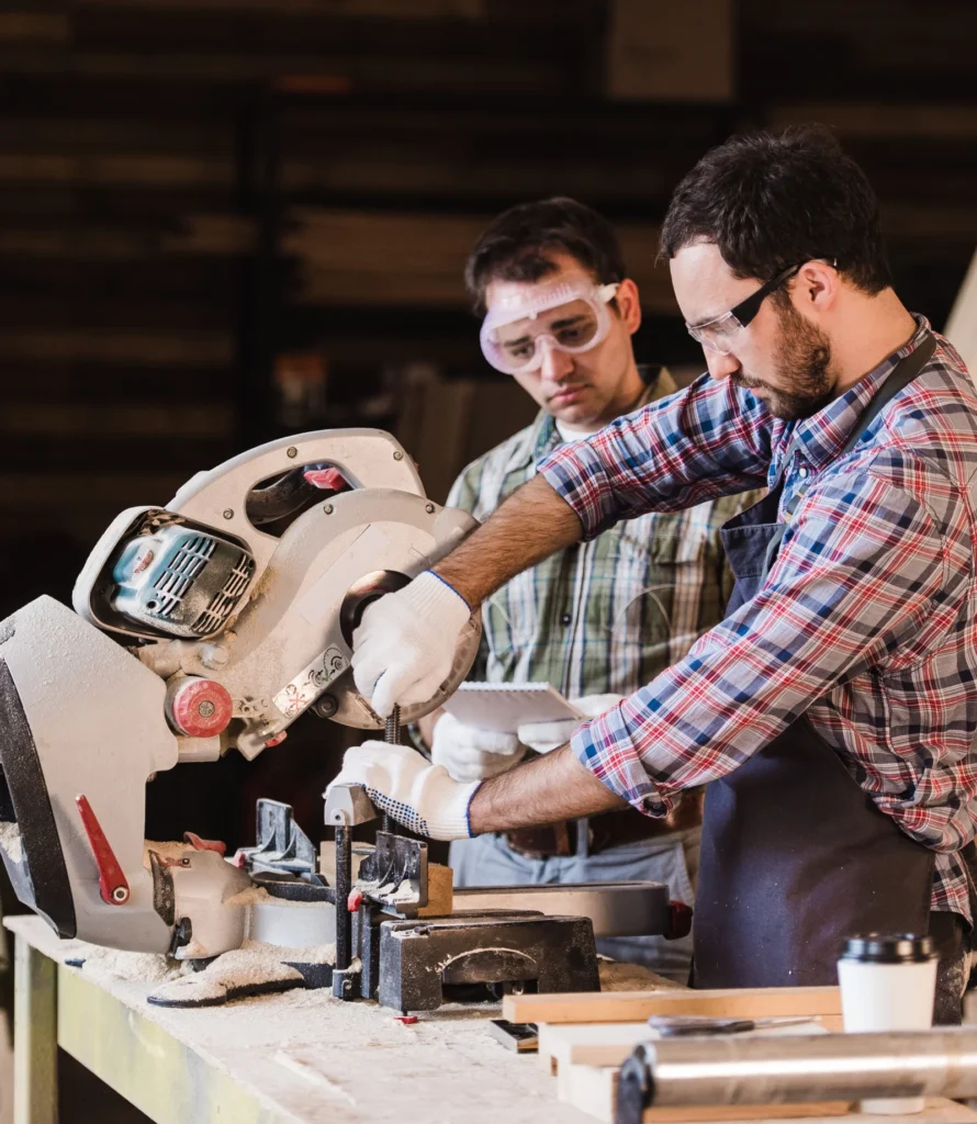 Stiligt - Två män i skyddsglasögon och arbetskläder använder en cirkelsåg för att kapa trä på en arbetsbänk i en verkstad. Fördjupning: Snickarens roll i skapandet av ett platsbyggt kök - expertens guide