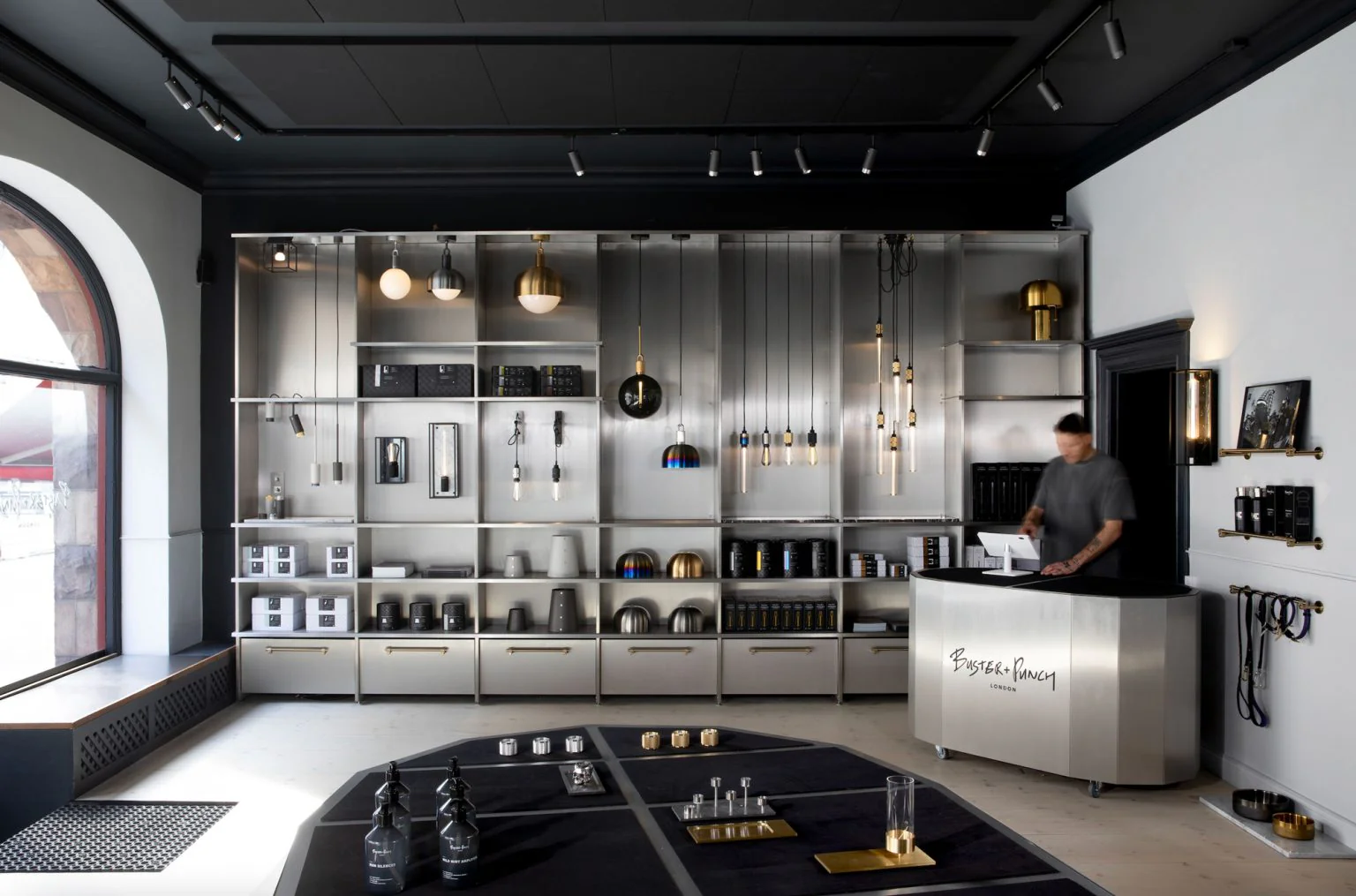 Stiligt - En modern butiksinredning med metallhyllor som visar olika hemvaror och belysningsarmaturer; en person står bakom en disk märkt "Buster + Punch".