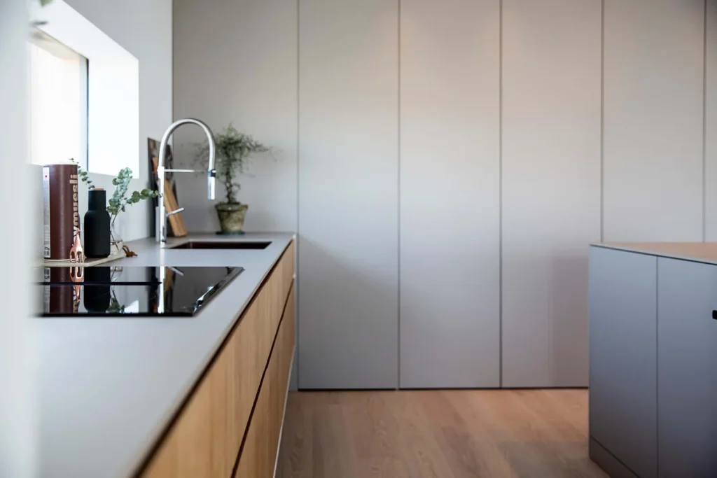 Stiligt - Modernt kök med skåp i ljust trä, snygga grå bänkskivor, inbyggd spishäll och minimalistisk inredning. Ett platsbyggt kök billigare på sikt ger mervärde, medan en krukväxt ger liv nära fönstret. Varför ett platsbyggt kök ofta är billigare än du tror på sikt: Smarta spartips!