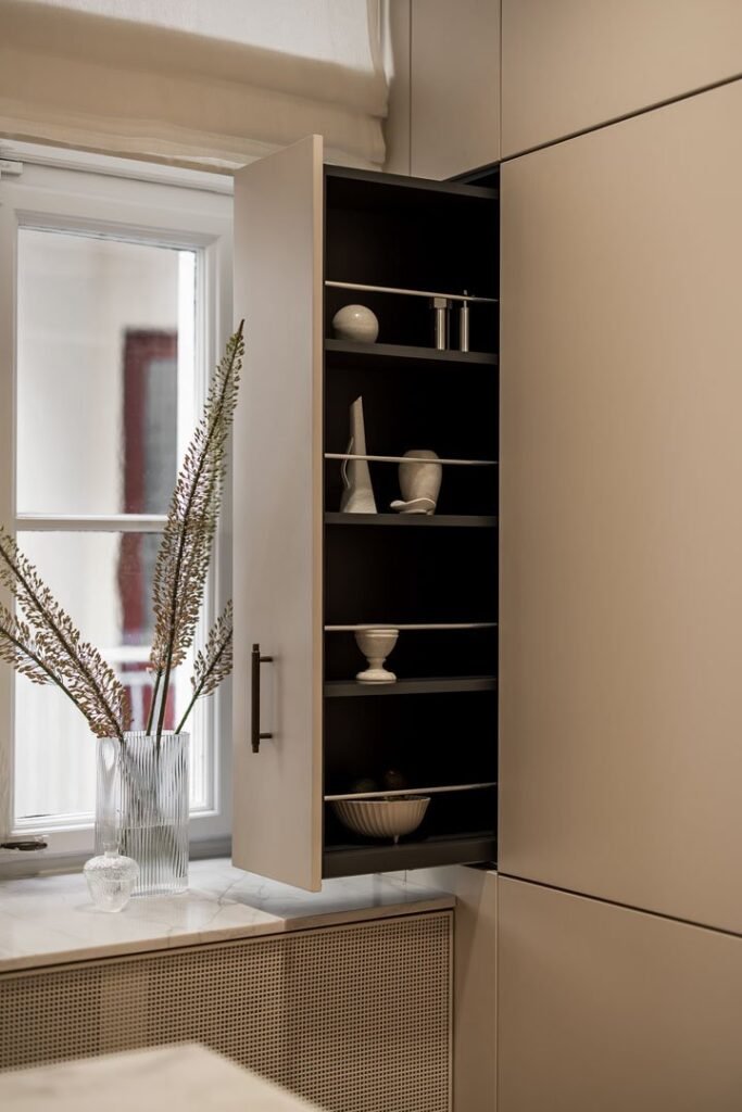 Stiligt - Ett modernt köksskåp med öppen dörr avslöjar hyllor med vita tallrikar och koppar; en glasvas med höga torkade växter står på bänken vid ett fönster. Sp planerar du optimal förvaring i ditt platsbyggda kök: smarta tips för maximal ordning