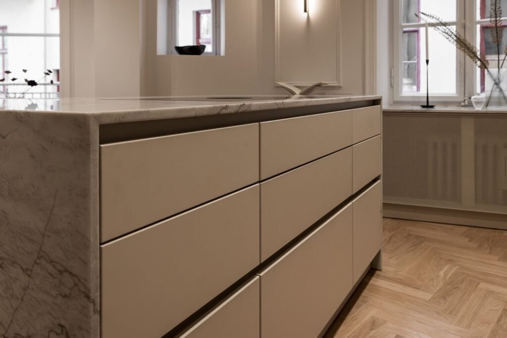 Stiligt - Minimalistisk köksö med beige lådor och en bänkskiva i marmor, placerad på ett fiskbensmönstrat trägolv i ett rum med stora fönster och neutral inredning. Så planerar du optimal förvaring i ditt platsbyggda kök: smarta tips för maximal ordning