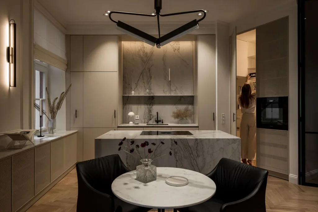 Stiligt - Modernt kök med marmorytor, runt matbord med svarta stolar i förgrunden och en person som står nära det öppna skafferiet i bakgrunden. Ett platsbyggt skafferi kan vara den gömda lyxen som förändrar allt.