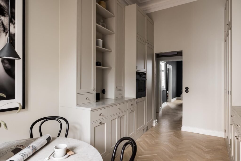 Stiligt - Modernt kök med ljusbeiga skåp, bänkskiva i marmor, runt bord med två svarta stolar och fiskbensmönstrat trägolv. En tidning och en kaffekopp står på bordet. Så skapade en lägenhetsägare sitt egna sekelskifteskök i City