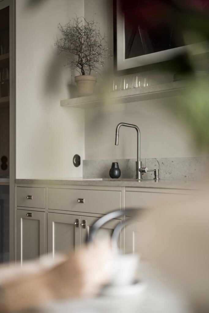 Stiligt - Minimalistiskt kök med ljusa skåp, en bänkskiva i marmor, en modern kran, en krukväxt på en hylla och suddiga föremål i förgrunden.Så förbereder du rummet inför installation: experttips för smidig start