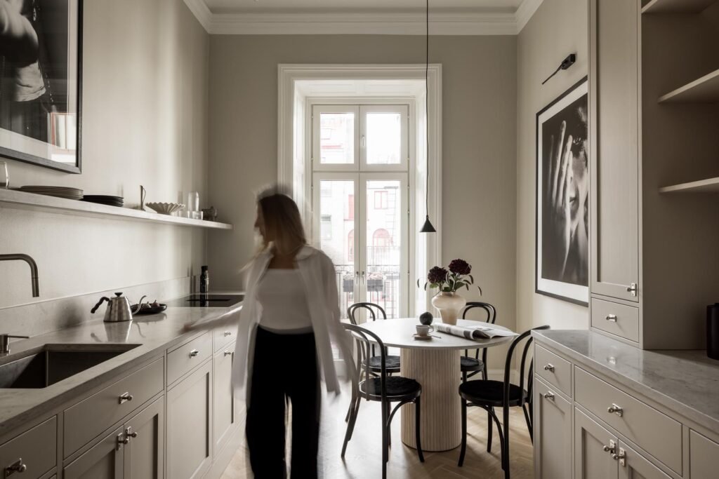 Stiligt - En kvinna står i ett modernt kök med beige skåpinredning, bänkskivor i marmor och en liten matplats vid ett stort fönster med naturligt ljusinsläpp. Kunder berättar: varför vi valde platsbyggt istället för ikea - bättre kvalitet