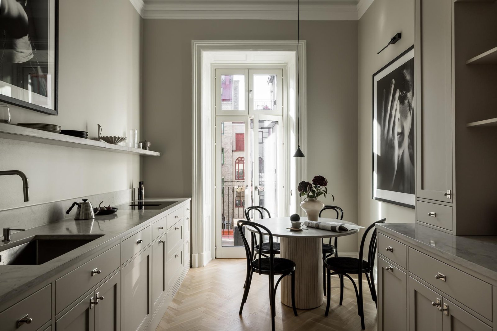 Stiligt - Ett modernt kök med ljusgrå skåp, bänkskivor i marmor, ett runt matbord med svarta stolar och inramad konst på väggarna, med utsikt över en balkong genom höga glasdörrar. Så skapade en lägenhetsägare sitt egna sekelskifteskök i City