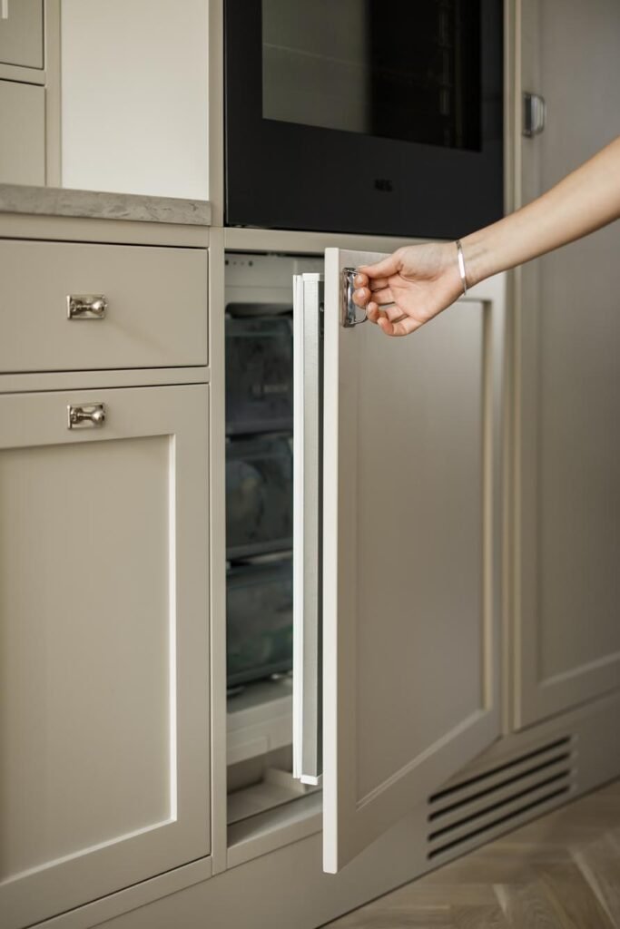 Stiligt - En persons hand öppnar en inbyggd fryslåda under en köksbänk med beige koksdesign-goteborg skåp. Köksdesign Göteborg: inspirerande ideer för ditt drömkök