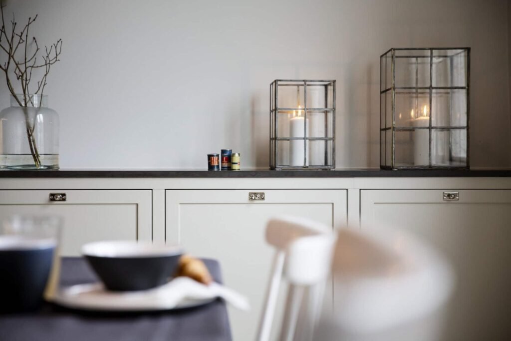 Stiligt - En minimalistisk inredning med vita skåp, två glaslyktor med tända ljus, burkar på bänkskivan och en bar kvist i en glasvas. Utrymmet utstrålar naturlig rörelse och balanserad köksdesign, med ett bord med skålar i förgrunden. Hur du skapar naturlig rörelse och balans i köksdesignen: Smarta tips!