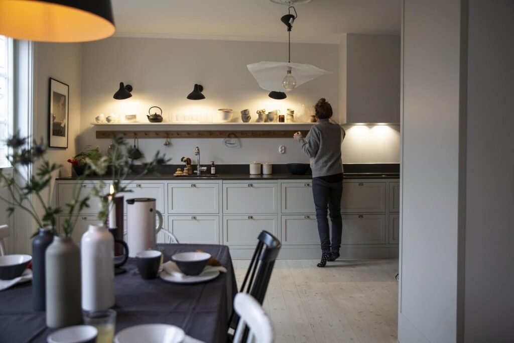 Stiligt - En person står vid en ljusgrå köksbänk och ordnar saker på en hylla, med ett matbord i förgrunden - vilket framhäver naturlig rörelse och balanserad köksdesign i utrymmet. Hur du skapar naturlig rörelse och balans i köksdesignen: Smarta tips!