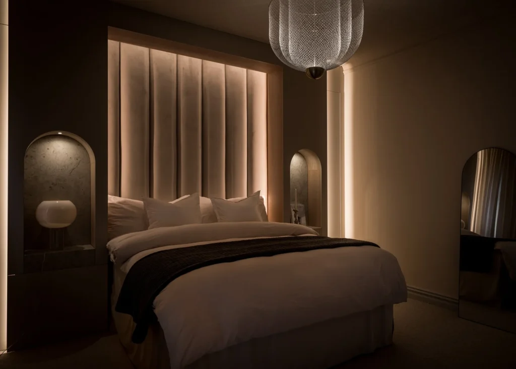 Stiligt - Ett modernt sovrum med en stor säng, dämpad belysning, vertikal vadderad sänggavel, inbyggda sänghyllor och en hängande armatur i nät.