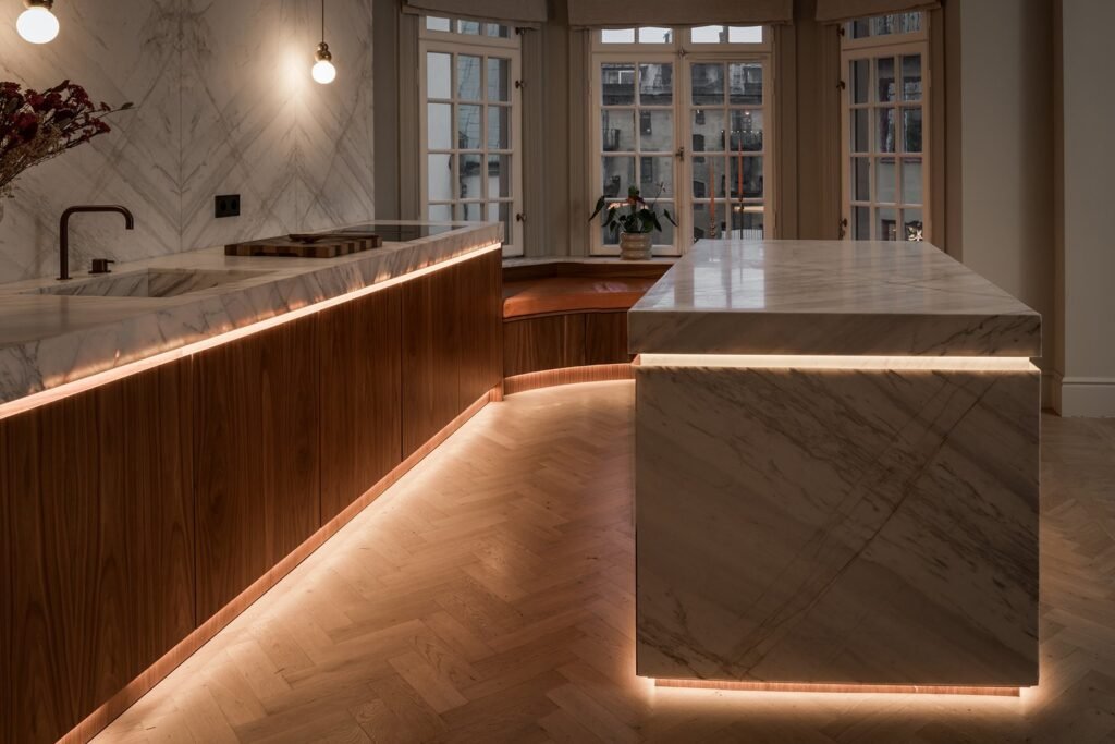 Stiligt - Modernt kök med bänkskivor i marmor, skåp i trä, fiskbensmönstrat trägolv och varm LED-belysning, med stora fönster i bakgrunden. Snyggt kök i trä och marmor: Stilrent & tidlöst designinspiration