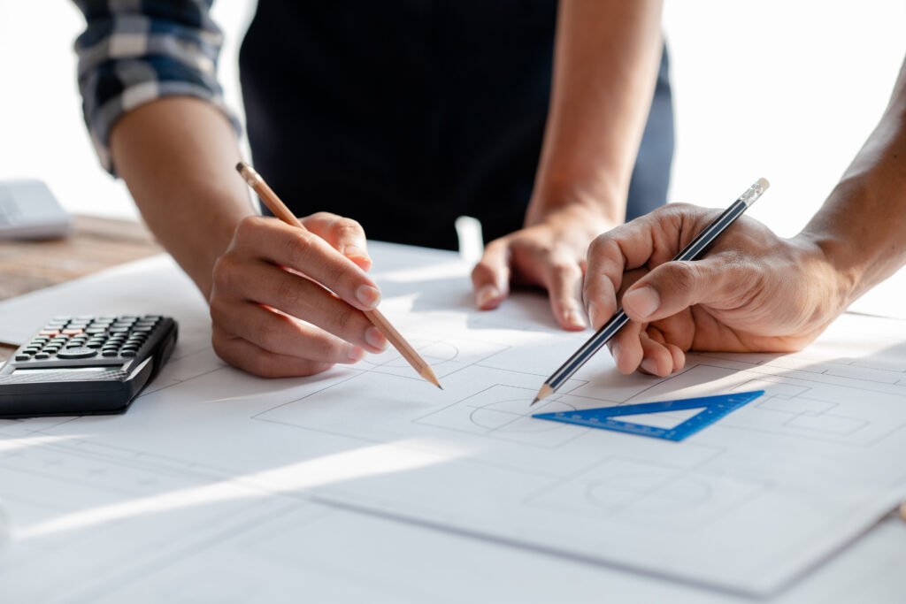 Stiligt - Två personer arbetar med arkitektritningar med pennor och linjal på ett bord, tillsammans med en miniräknare och grafpapper. Planlösningens betydelse för trivsel i köket: Skapa harmoni & funktion