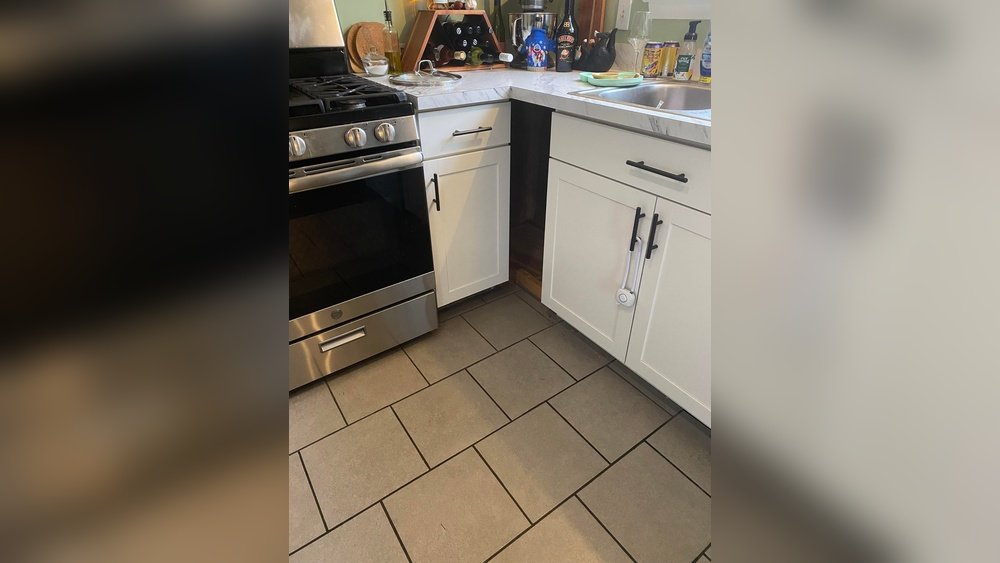 Stiligt - Kökshörna med spis i rostfritt stål, vita skåp, bänkskiva med olika föremål och diskho, golvet är kaklat med stora grå rutor.