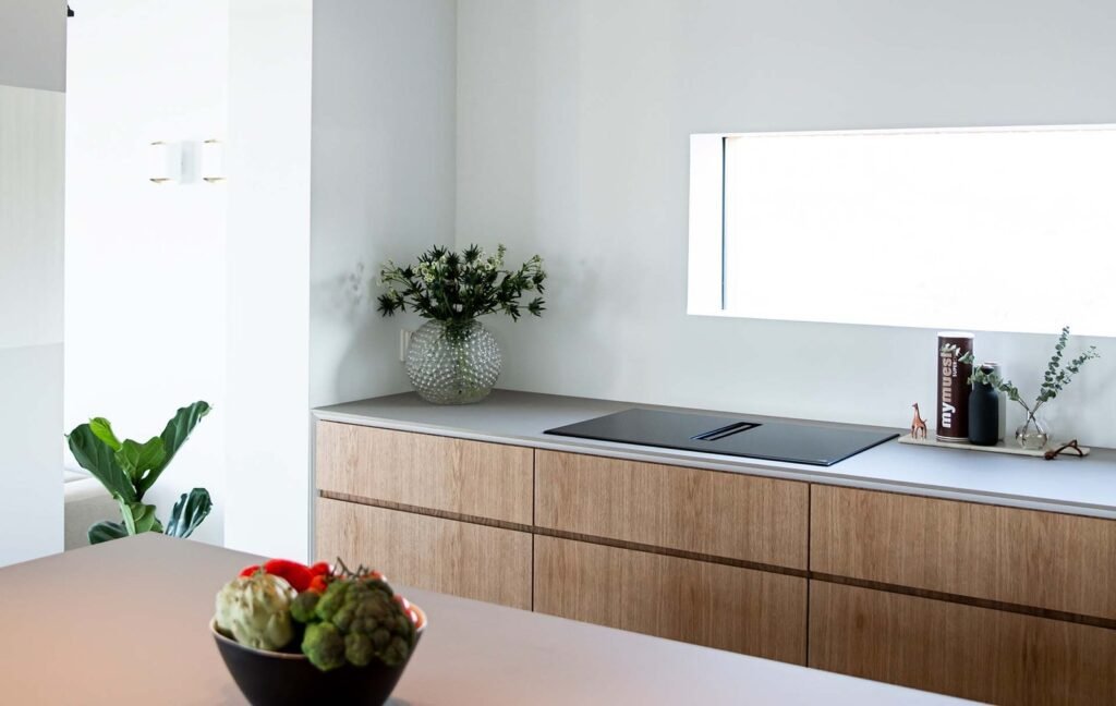 Stiligt - Ett modernt kök med skåp i ljust trä, en inbyggd spishäll, en vas med blommor, böcker och en skål med grönsaker på bänken. Ett fönster ger naturligt ljus.Köksdesign för barnfamiljer - funktion möter vardag: Smart & stilrent!