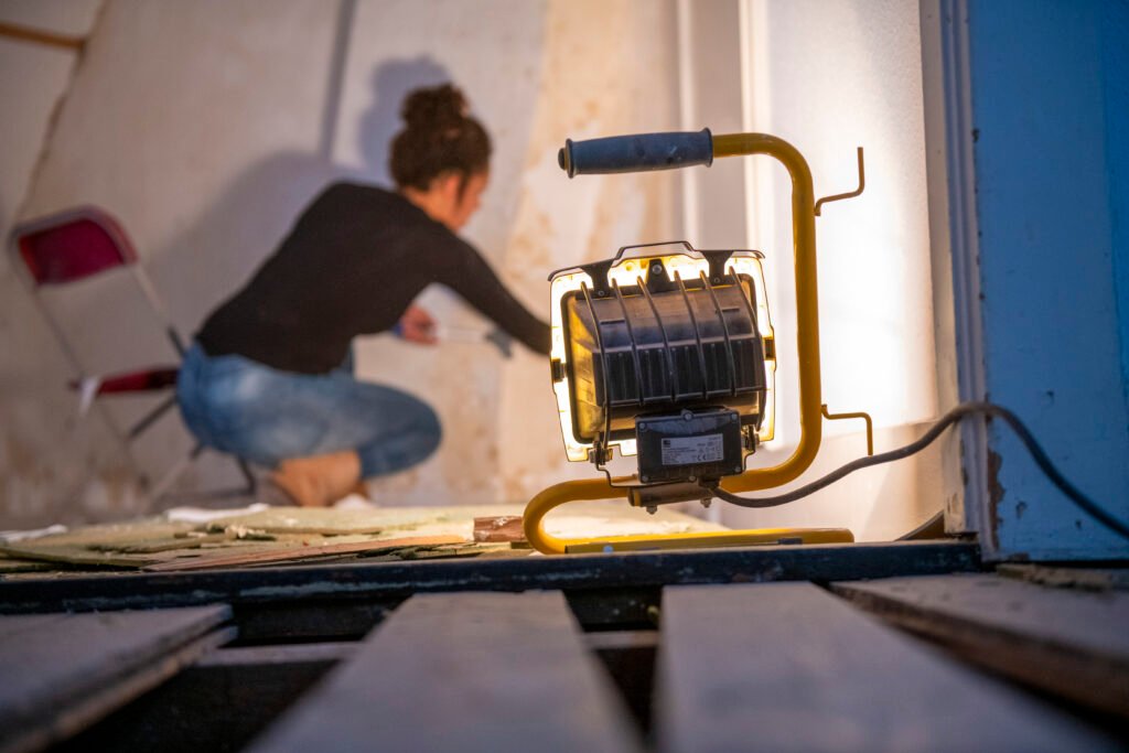 Stiligt - En arbetslampa lyser upp ett rum där en person i fritidskläder sitter på huk och arbetar på en vägg i närheten av en röd fällstol. Så förbereder du rummet inför installation: experttipsen för smidig start!