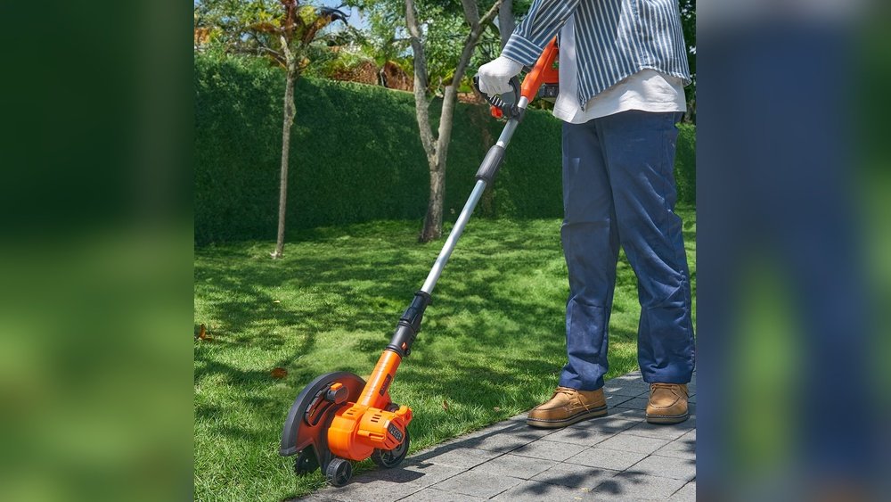 Stiligt - Person som använder en orange elektrisk grästrimmer för att klippa gräsmattan intill en asfalterad gångväg i en trädgård.