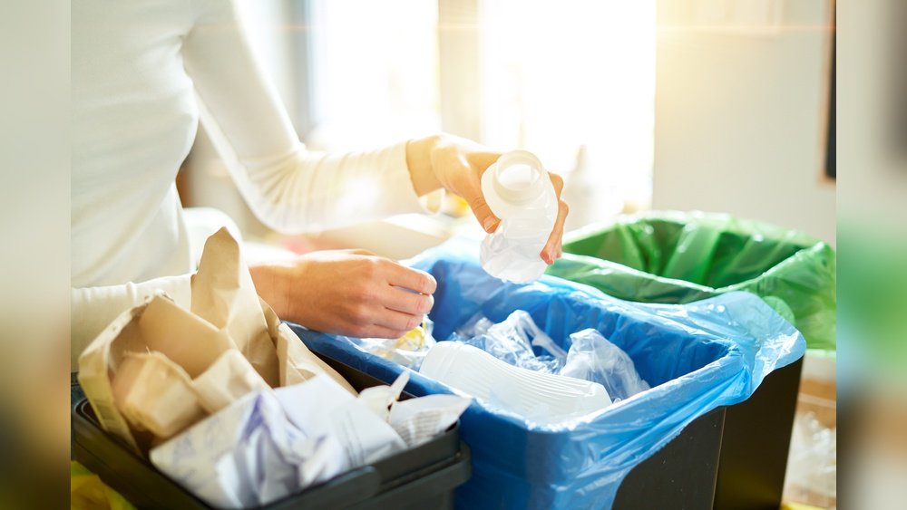 Stiligt - En person sorterar återvinningsbart material och lägger en plastflaska i en blå behållare, med papper och andra återvinningskärl synliga i närheten.