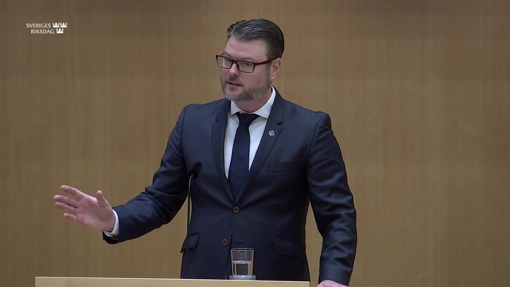Stiligt - En man i kostym och slips talar vid ett podium med ett glas vatten framför sig, i en formell miljö med texten "Sveriges Riksdag" på väggen.
