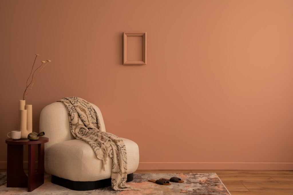 Stiligt - En cremefärgad fåtölj med en mönstrad pläd står bredvid ett litet sidobord med vaser mot en persikofärgad vägg med en tom ram. En matta och ett trägolv kompletterar miljön. Inredning varma färger