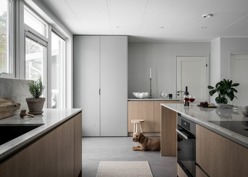 Stiligt - Modernt kök med skåp i ljust trä, en köksö och stora fönster; en hund ligger på golvet nära en liten pall och en växt står på bänken. De vackraste kulörerna för platsbyggda kök just nu: Trendiga färger 2025