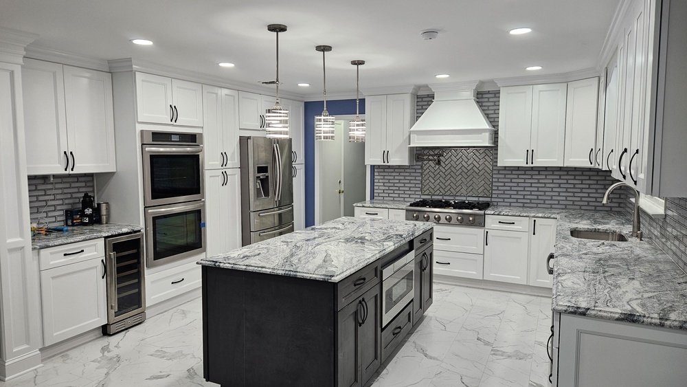 Stiligt - Modernt kök med vita skåp, rostfria vitvaror, grå kakelplattor, bänkskivor i marmor, en ö med inbyggd ugn och pendellampor.