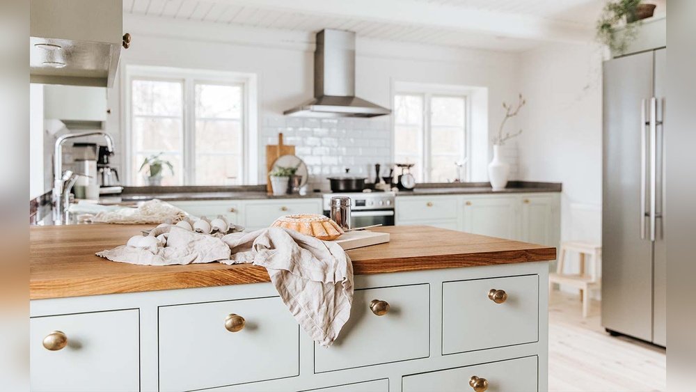 Stiligt - Modernt kök med ljusgröna skåp, bänkskiva i trä, vitvaror i rostfritt stål och en limpa bröd på en trasa i förgrunden. Naturligt ljus flödar in genom två fönster.