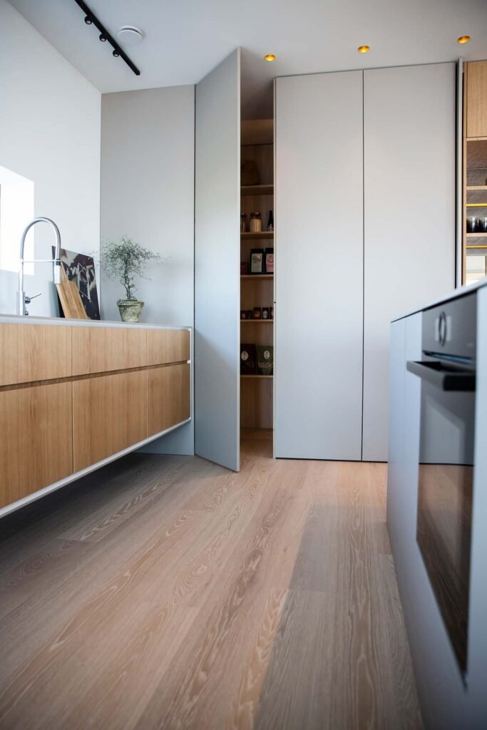 Stiligt - Modernt kök med ljust trägolv, minimalistiska skåp, en delvis öppen skafferidörr som avslöjar hyllor med matvaror och en liten krukväxt på arbetsbänken. Köksdesign för barnfamiljer - funktion möter vardag: Smart & stilrent!