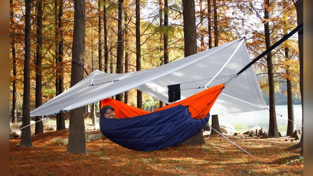 Stiligt - Person liggande i en hängmatta med en presenning över sig, uppställd bland höga träd i en skog med höstlöv på marken.