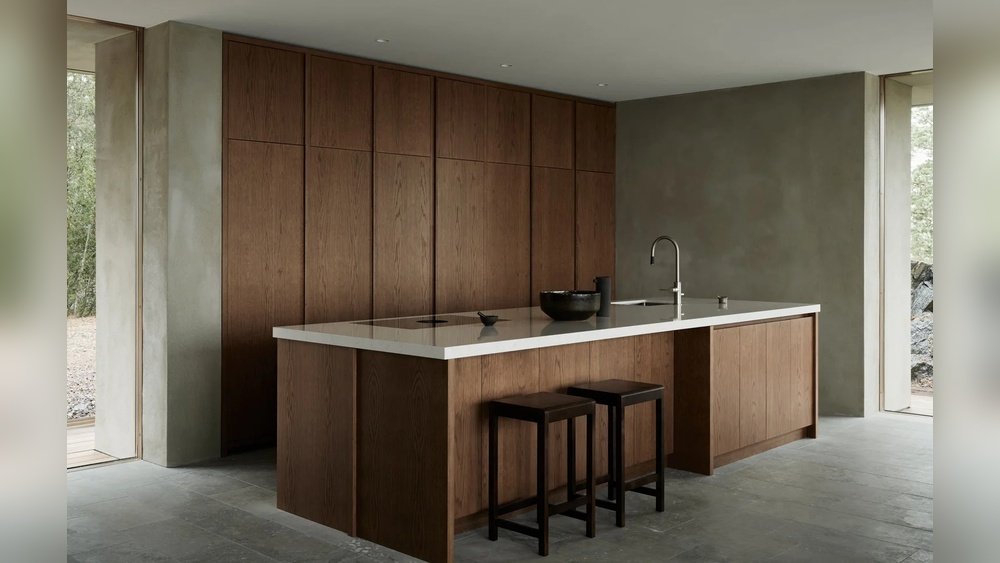 Stiligt - Minimalistiskt kök med bruna träskåp, en vit bänkskiva med integrerad diskho, två bruna pallar och neutrala grå väggar och golv.