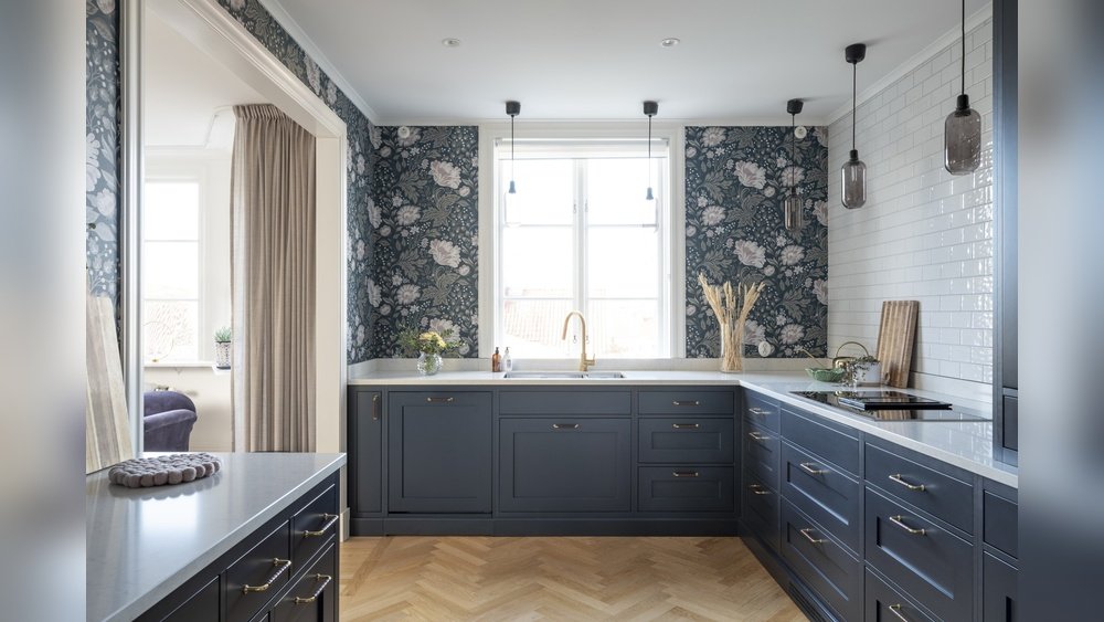 Stiligt - Modernt kök med blå skåp, blommig tapet, vitt kakel, pendellampor och ett stort fönster ovanför diskbänken.