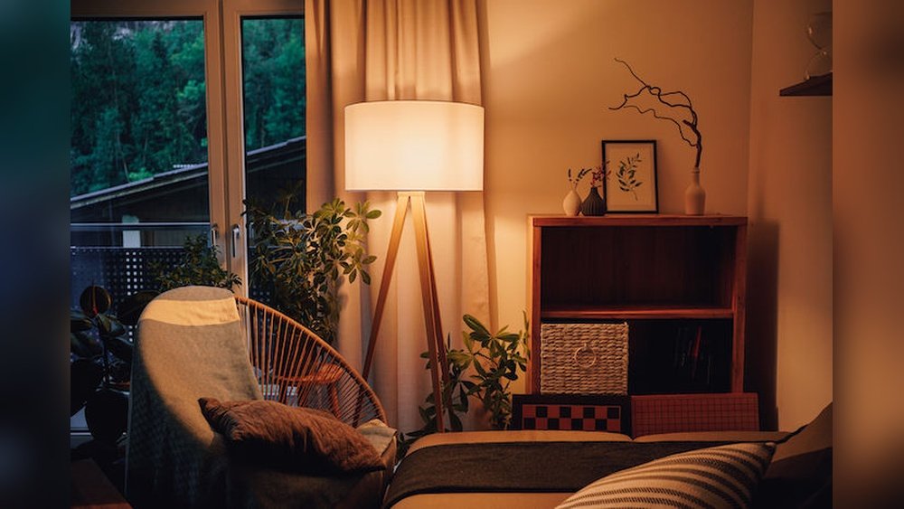 Stiligt - Mysigt vardagsrum med golvlampa, fåtölj, bokhylla, krukväxt och ett fönster som visar grönska utanför. Varm belysning skapar en avslappnad atmosfär.