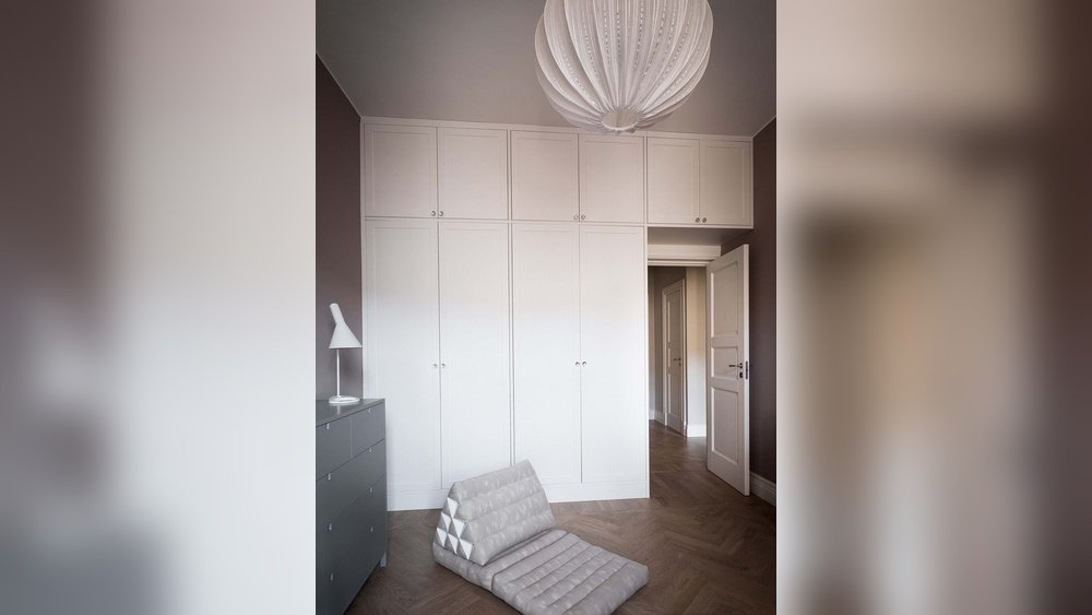 Stiligt - Ett minimalistiskt rum med vita inbyggda skåp, en öppen dörr, en grå byrå med en lampa, en modern taklampa och en hopfälld grå golvstol på ett fiskbensmönstrat trägolv.