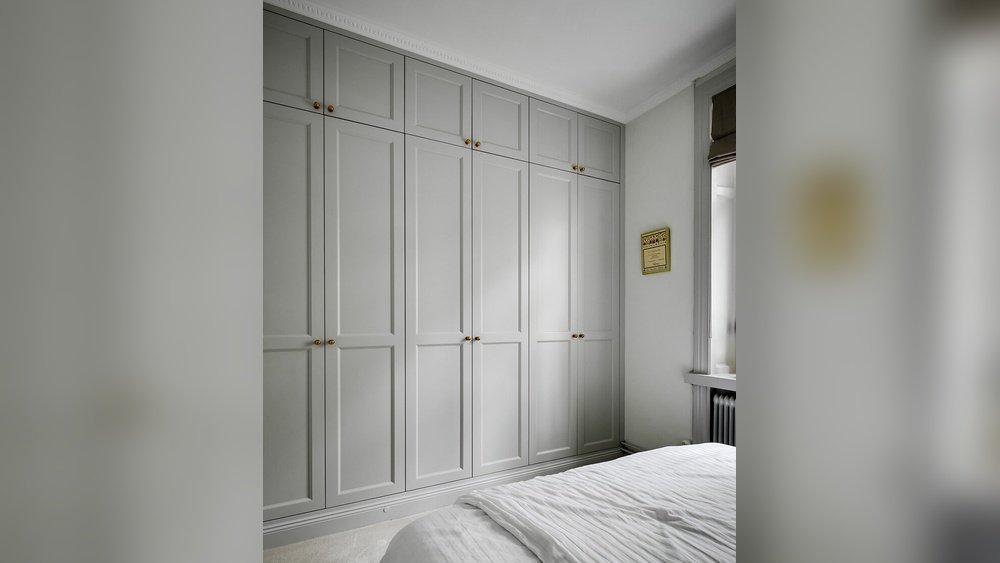 Stiligt - Inbyggda grå garderober från golv till tak med mässingshandtag i ett sovrum med vita sängkläder och ett fönster med en roman shade.