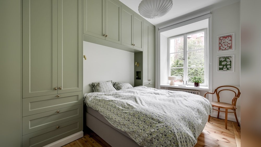 Stiligt - Ett litet sovrum med en dubbelsäng, gröna inbyggda skåp, en trästol, inramad konst på väggen och ett fönster som släpper in naturligt ljus.