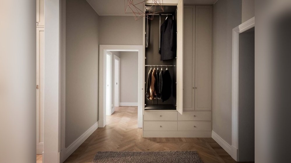 Stiligt - En minimalistisk, inbyggd garderob med öppna dörrar visar hängande kläder och lådor i en neutral hall med fiskbensmönstrat trägolv.