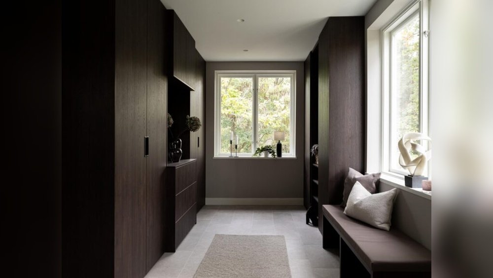 Stiligt - En modern hall med skåp i mörkt trä, en bänk med kuddar och ett fönster som släpper in naturligt ljus. En neutral matta ligger på det ljusa klinkergolvet. Skulpturer och växter dekorerar utrymmet.