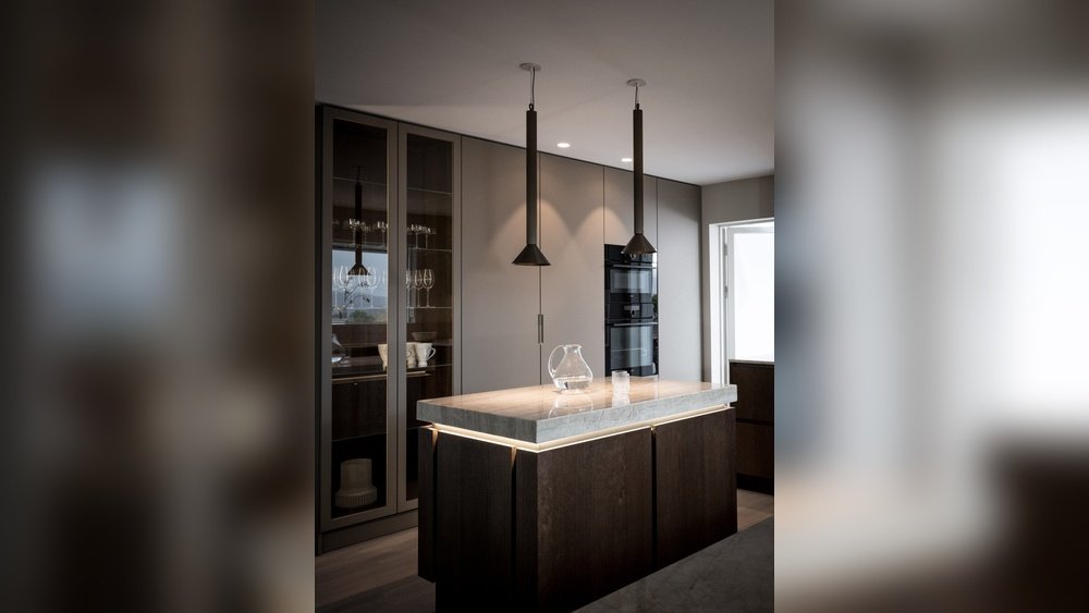 Stiligt - Modernt kök med en central köksö, två pendellampor, skåp med glasfasad, inbyggda ugnar och en glaskanna på bänkskivan.