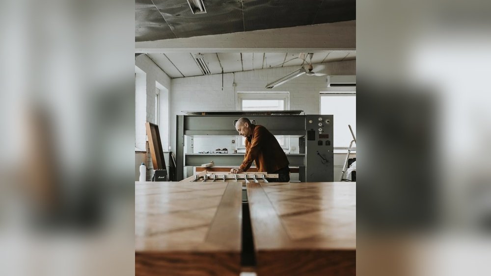 Stiligt - En person arbetar med ett träprojekt i en verkstad, stående bakom ett stort bord med träbitar och verktyg synliga.