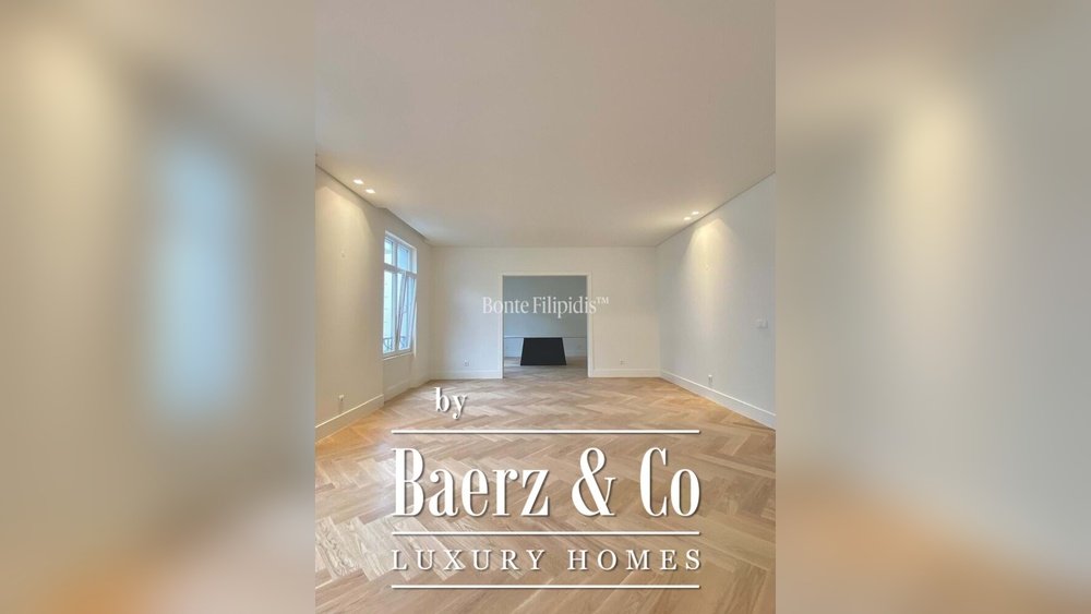 Stiligt - Rymligt, tomt vardagsrum med fiskbensparkett, vita väggar, infälld belysning, stort fönster och en central svart öppen spis. Textöverlägget lyder "Baerz & Co Luxury Homes.